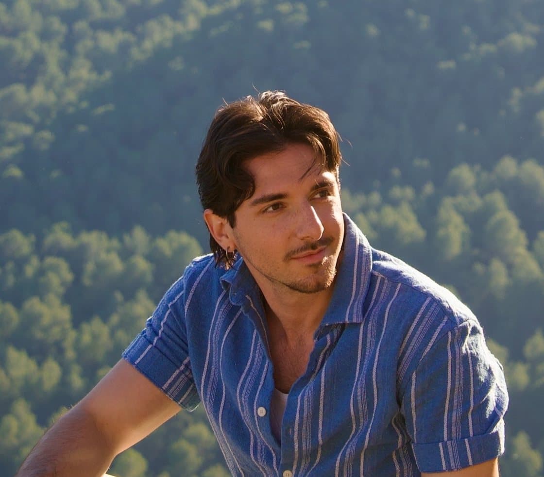 Portrait of Arvin Ansari seated outdoors in a blue striped shirt, warm sunlight and a green forest backdrop.